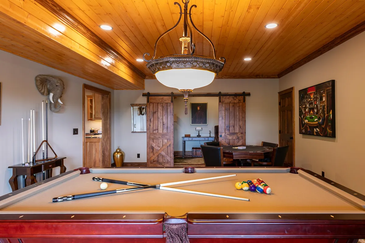 A spacious game room with a pool table at the center, illuminated by a chandelier, with wooden sliding doors, an elephant wall sculpture, and a view into a dining area. The room effortlessly blends spaces for entertainment and social gatherings.