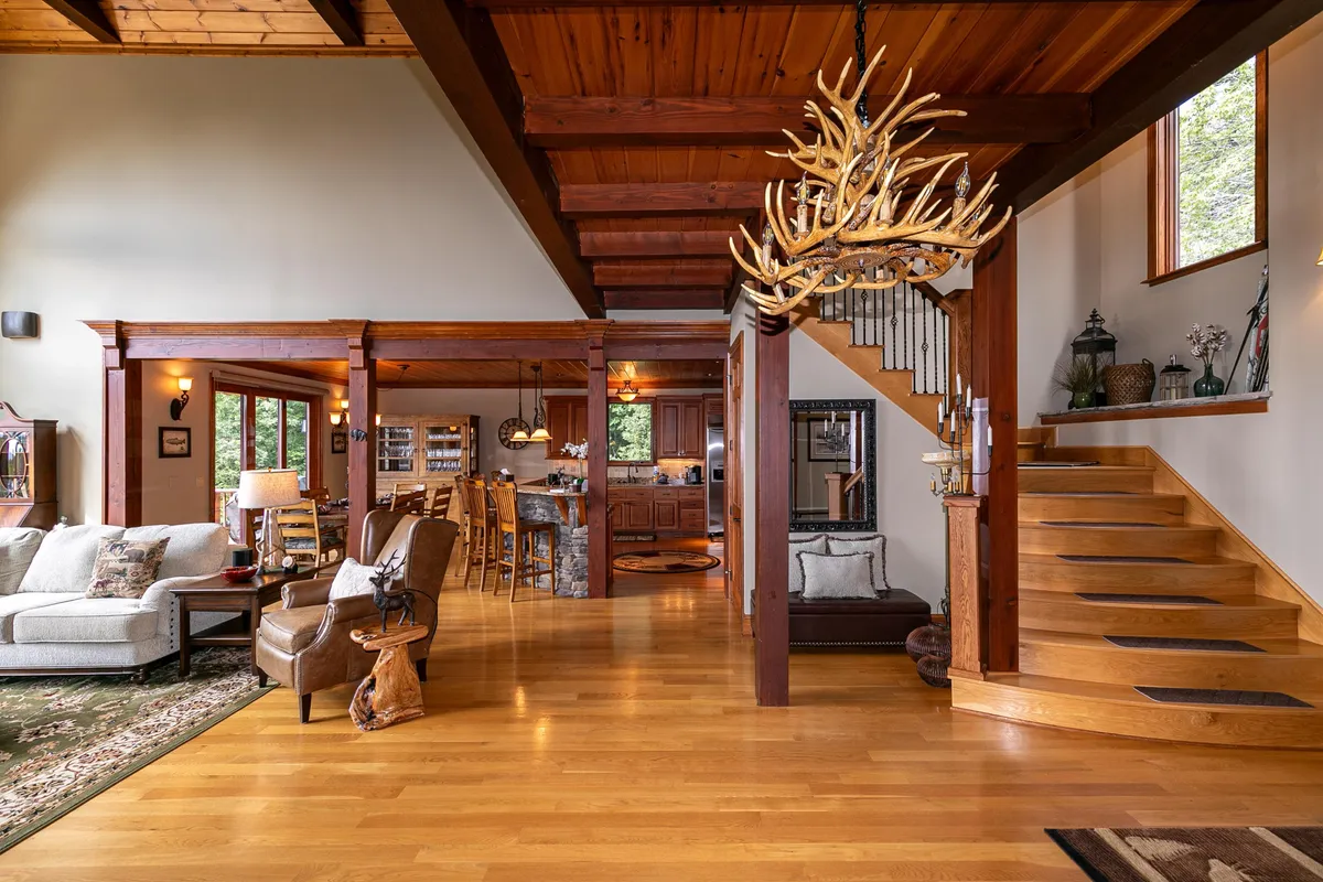 The foyer area transitions into a cozy sitting space, anchored by a dramatic antler chandelier and a beautifully crafted wooden staircase. Comfortable seating invites guests to linger and connect, while glimpses of the kitchen further emphasize the open flow between communal spaces.