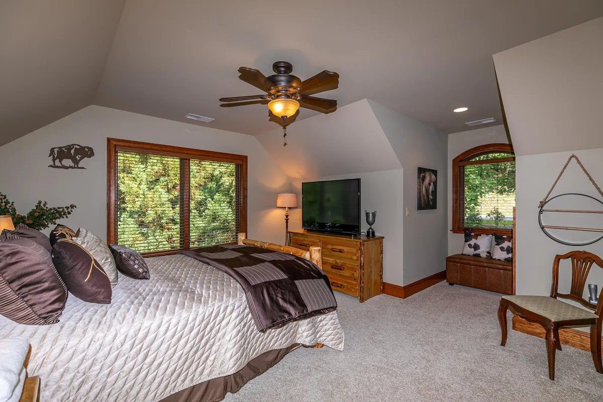 A serene bedroom featuring a well-made bed adorned with decorative pillows, elegant wooden furniture, large windows welcoming in natural light, and a ceiling fan overhead. The setting exudes tranquility, offering views of surrounding nature—a perfect spot for unwinding with a book or enjoying a peaceful morning.