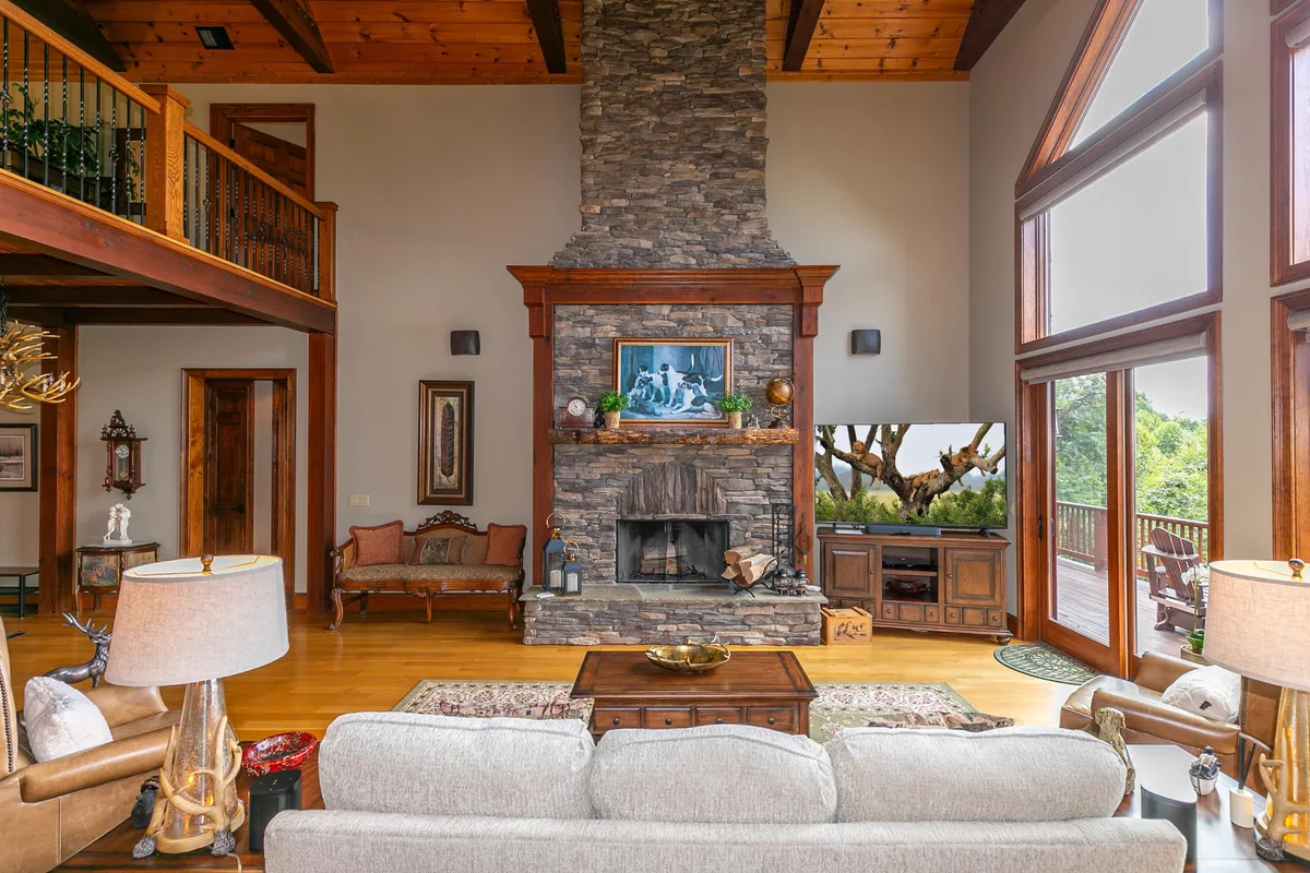 A sophisticated living area centered around a rustic stone fireplace, with large windows framing the lush landscape outside. A plush sofa and curated artwork create a refined yet comfortable ambiance ideal for both relaxation and entertaining.