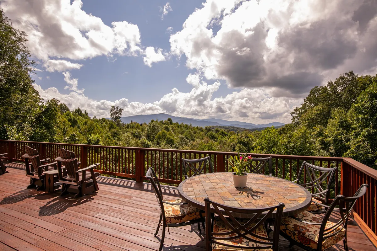 A spacious wooden deck with a round table and chairs overlooks a breathtaking panoramic mountain view under a cloudy sky. The scene suggests tranquility and the opportunity for outdoor dining, relaxing, or social gatherings surrounded by nature.