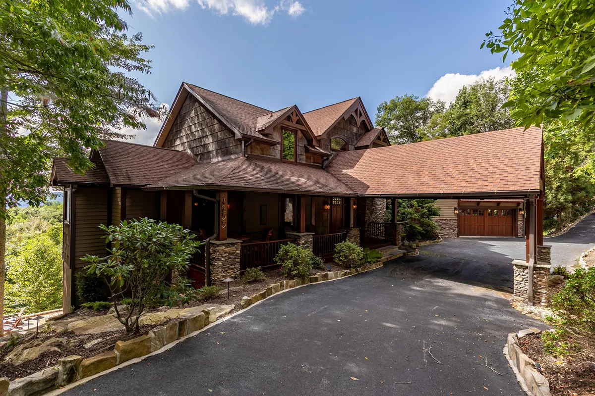 The image showcases the striking architecture of a luxury single-family mountain home surrounded by lush greenery, with a stone pathway leading guests toward a welcoming entrance. The generous porch invites relaxation and offers panoramic views of the Blue Ridge Mountains, while manicured gardens hint at tranquil outdoor spaces for gathering and exploration.