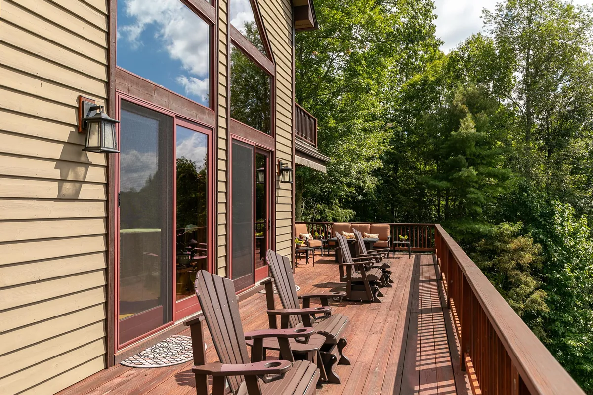 An inviting wooden deck features Adirondack chairs and large glass windows, framed by lush greenery. The setting is perfect for relaxing, entertaining guests, or enjoying the peaceful mountain atmosphere.