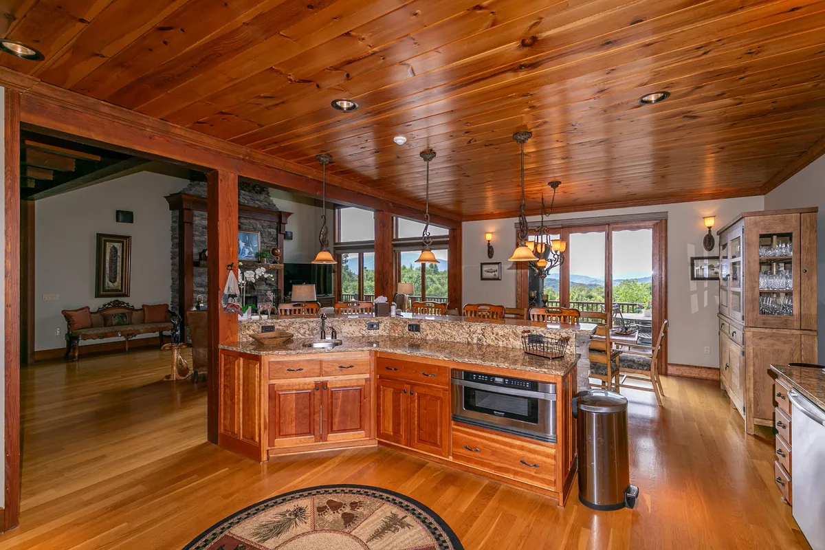 A spacious kitchen with a large island takes center stage, surrounded by rich wood cabinetry and gleaming granite countertops. Stainless steel appliances line the walls, and expansive windows behind the dining area offer striking views of the forested landscape. The open-plan design makes it easy to move between the kitchen and the dining area, encouraging communal cooking and shared meals.