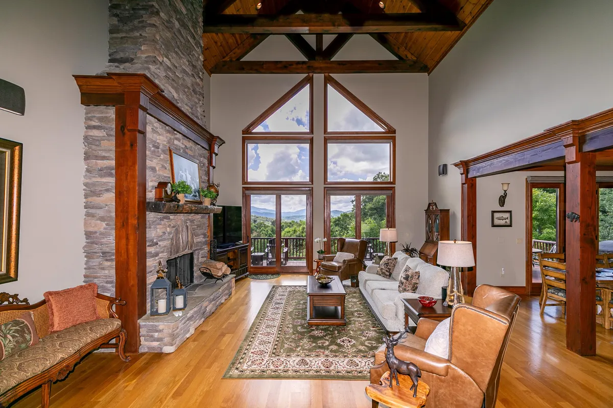A welcoming open-concept living area featuring a striking stone fireplace, exposed wooden beams, a comfortable seating arrangement, and expansive windows that flood the space with natural light and offer scenic views of the Blue Ridge Mountains. An elegant decorative rug and thoughtfully arranged furniture further enhance the sense of warmth and social connection.