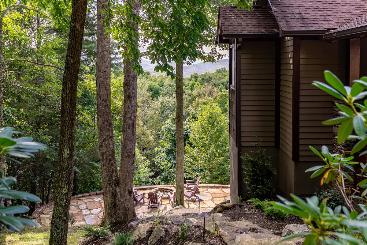 A serene stone patio with wooden chairs facing distant, rolling mountains and ringed by trees. The setting is ideal for outdoor dining, social gatherings, or quiet reflection, seamlessly integrating with the natural environment.